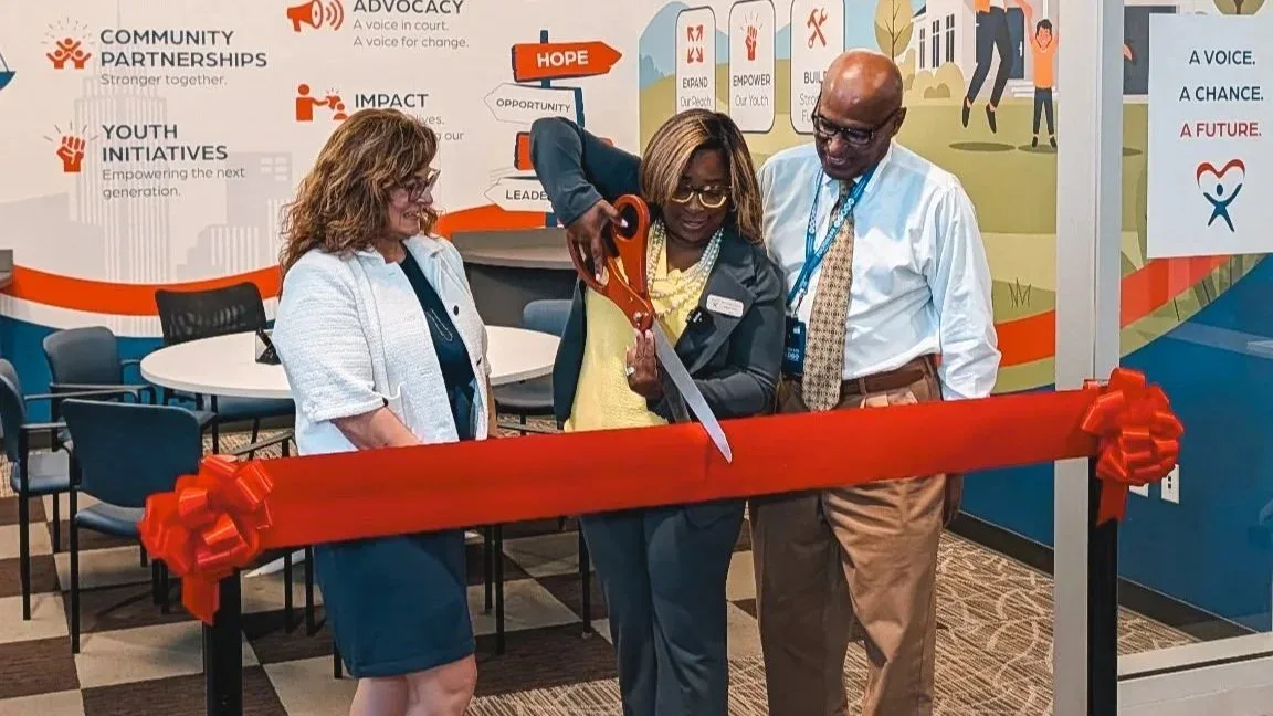 A woman cuts a red ribbon with oversized scissors, flanked by a man and a woman, in front of a wall listing community partnerships, youth initiatives and impact