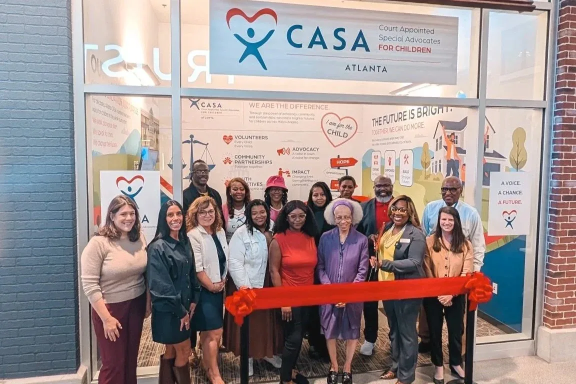 About 15 people stand behind a red ribbon outside the Atlanta CASA storefront under a large CASA Court Appointed Special Advocates for Children sign