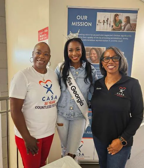 Three women pose in front of an Atlanta CASA mission banner, one wearing a CASA 30 Years T-shirt, one wearing a crown and a Miss Georgia sash