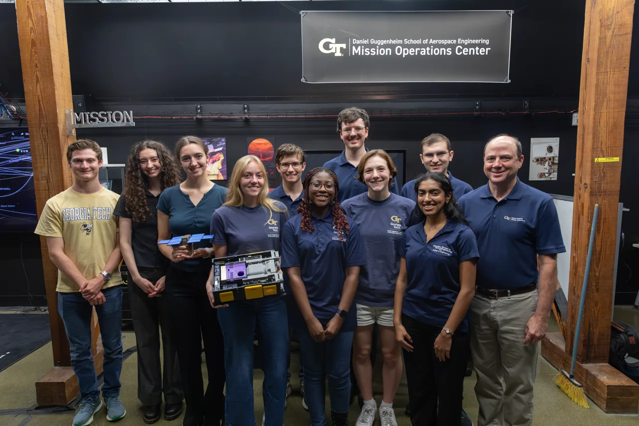 Georgia Tech students, faculty and staff, including professor Glenn Lightsey, stand under a sign reading Daniel Guggenheim School of Aerospace Engineering Mission Operations Center, one holding spacecraft hardware